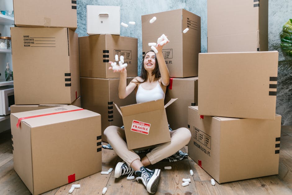 A young woman with long dark hair sitting cross-legged on a wooden floor surrounded by multiple large cardboard moving boxes, some sealed with red tape and others open, revealing packing materials such as foam packing peanuts which she is actively tossing into the air. She is smiling and looking upwards, inside a room with grey textured walls and a light-colored ceiling. The scene depicts a home relocation process with boxes ready for transport, supported by professional packing and moving services from Belsize Park Movers. Visible in the background are a small white shelf with a plant and other household items, indicating a domestic environment. The photograph captures the packing and loading phase of a house removal, emphasizing careful handling and organisation of belongings, relevant to furniture transport and relocation logistics.