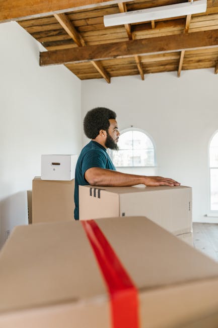 A man with dark curly hair and a beard, wearing a blue t-shirt, stands inside a bright room with a wooden ceiling and exposed beams, surrounded by neatly stacked medium-sized cardboard boxes sealed with packing tape, some with visible tape strips. The boxes are positioned on the floor and are being prepared for home relocation or furniture transport by Belsize Park Movers. The room has white walls and large arched windows allowing natural light to illuminate the space. The man appears to be in the process of organizing or moving the boxes, which are likely packed for a house move. Visible equipment includes no trolleys or packing materials in this shot, but the boxes are arranged in a way consistent with loading or unloading procedures during residential removals services. This scene depicts a typical indoor packing or loading process as part of a professional moving service, with the focus on careful handling and preparation for relocation.