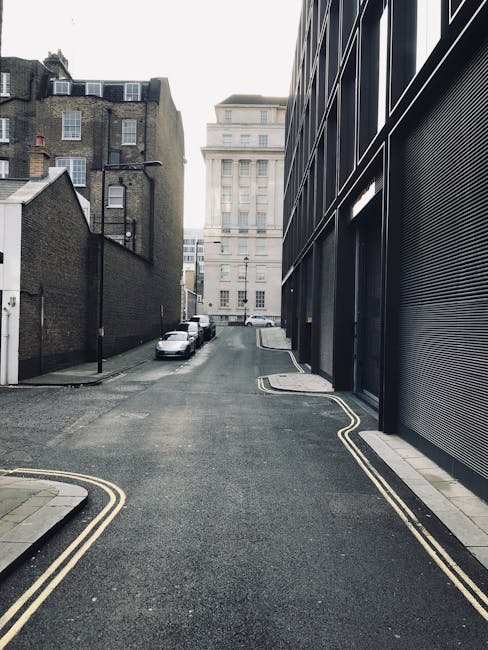 A narrow urban alleyway with a paved surface showing patches and markings, flanked by tall brick and metal-clad buildings on both sides. On the right, a closed black metal shutter with graffiti, and on the left, a metal fire escape ladder attached to the building's facade. Nearby, several cardboard boxes and protective packing materials are stacked on the ground. In the distance, a small black vehicle is parked near a doorway with signage, visible through the alley opening. The environment appears to be part of a home relocation process, with the setting prepared for furniture transport or loading, and ambient, diffuse lighting coming from above. The alley is used by Belsize Park Movers for house removals and moving logistics in tight city streets, emphasizing cautious navigation through confined spaces.
